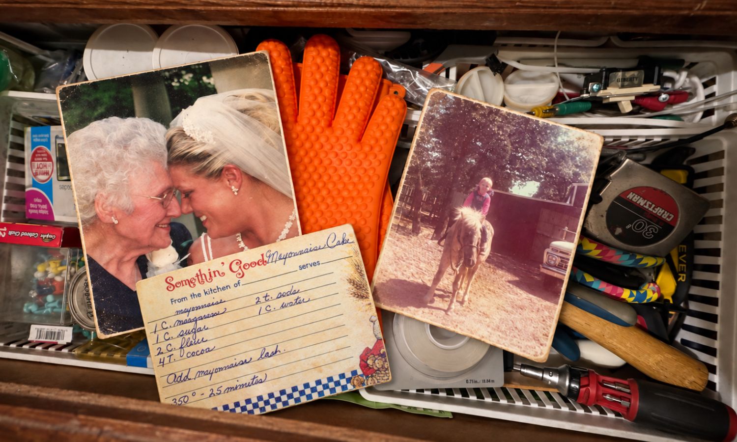 Open junk drawer filled with everyday tools and household clutter, with a wedding photo of the author and her grandmother, a childhood pony photo, and a handwritten family recipe card tucked among the items