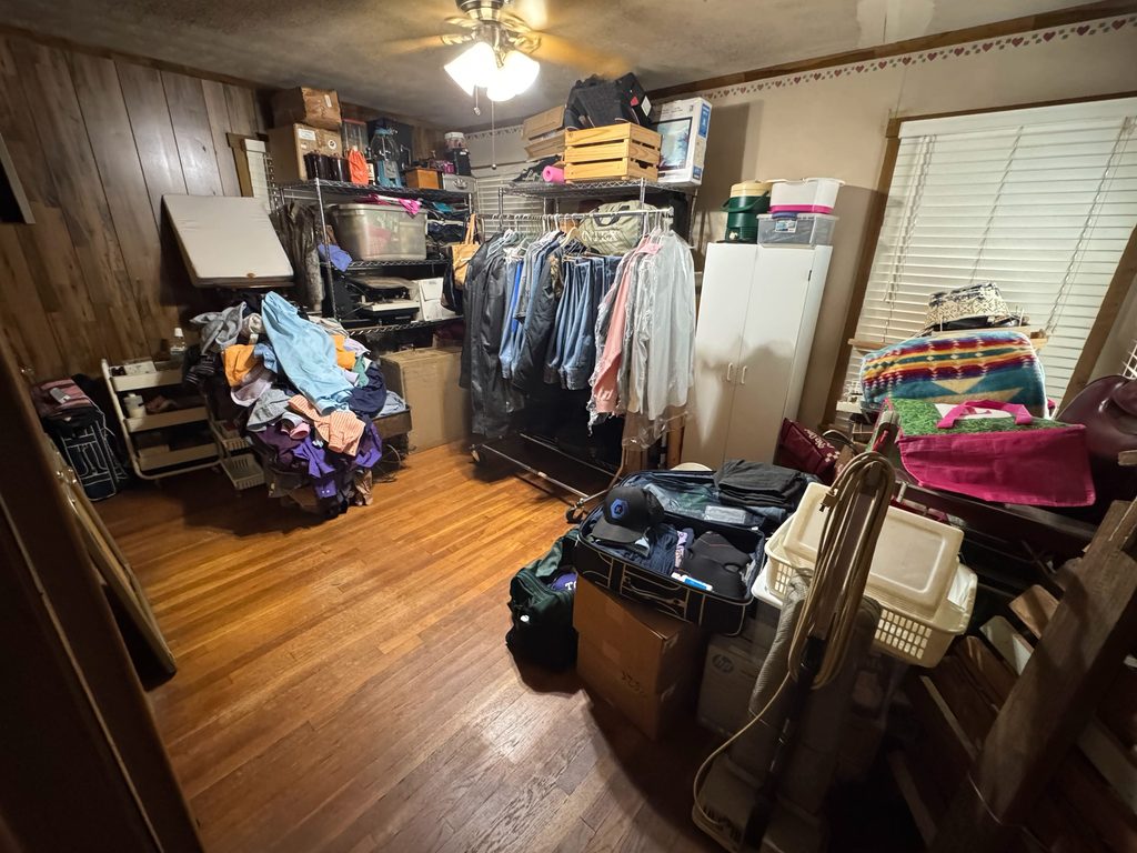 Cluttered room before a home office cosmetic upgrade, showing clothing racks, boxes, equipment, and storage waiting to be sorted.